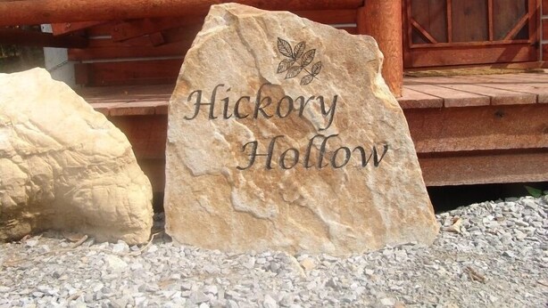 A stone sign with the text "Hickory Hollow" and a leaf design, placed in front of a wooden cabin with a porch. - The Hickory Hollow Log Cabin by Garden of Gods in the Shawnee National Forest (Elizabethtown)