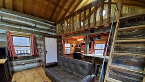 A cozy cabin interior with a sofa, wooden walls, a kitchen area, and a loft space.