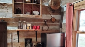A rustic kitchen with wooden walls, a stainless steel refrigerator, a wooden shelf with jars, and a hanging pot.
