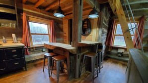A rustic cabin interior with a wooden dining table, two bar stools, a bar counter, and a window with curtains.