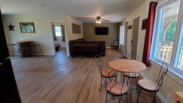 A spacious living area with a wooden floor, a round dining table with four chairs, a dark-colored sofa, and a ceiling fan.