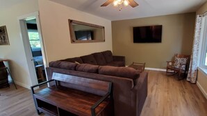A living room with a brown sofa, a wooden coffee table, a TV mounted on the wall, and a window with curtains.