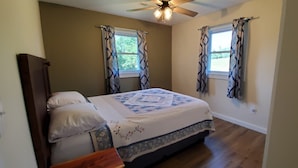 A bedroom with a bed, two windows with curtains, a ceiling fan, and wooden flooring.