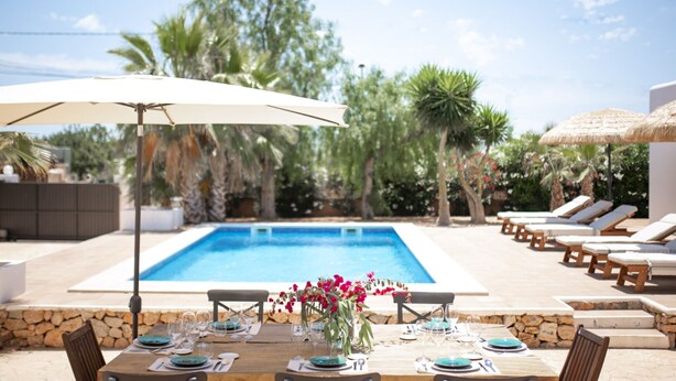 Een eetgedeelte aan het zwembad met een tafel voor twee personen, een zwembad, ligbedden en een rieten parasol.
