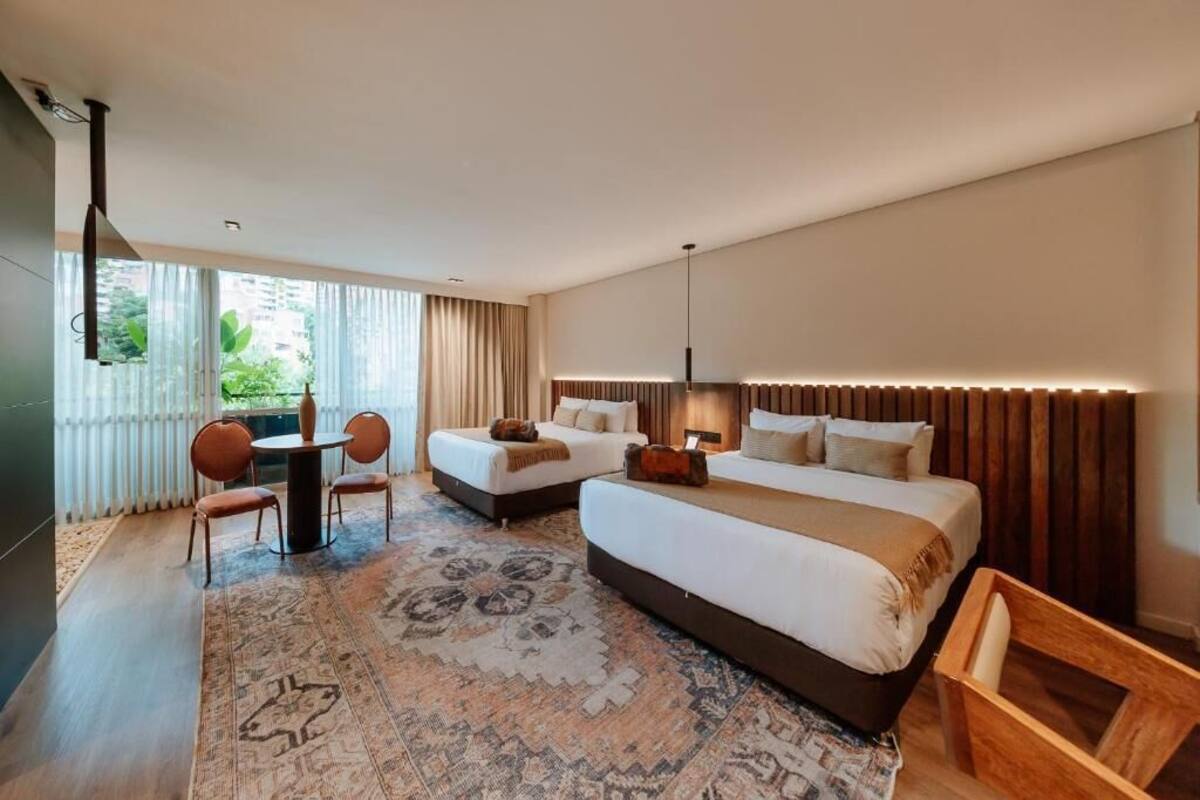 A modern hotel room with a large bed, a small table, two chairs, and a view of greenery outside.