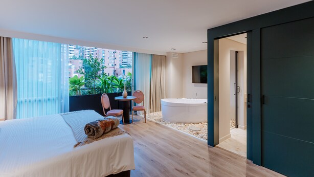 A modern hotel room with a large bed, a sitting area with a small table and chairs, a bathtub, and a view of the city through large windows. - 1616 HOTEL  (Medellín)