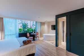 A modern hotel room with a large bed, a sitting area with a small table and chairs, a bathtub, and a view of the city through large windows. - 1616 HOTEL  (Medellín)