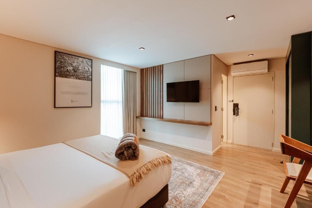 A modern hotel room with a bed, a flat-screen TV mounted on the wall, a wooden floor, and a large window with curtains.