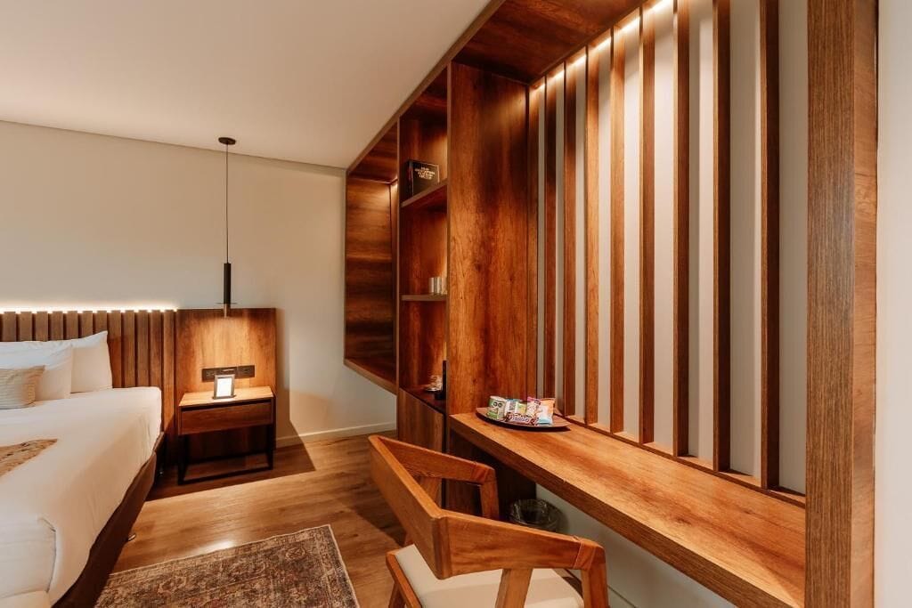 A modern hotel room with a wooden headboard, a bedside table with a lamp, a wooden bench, and a built-in shelf with books and a tray of snacks.