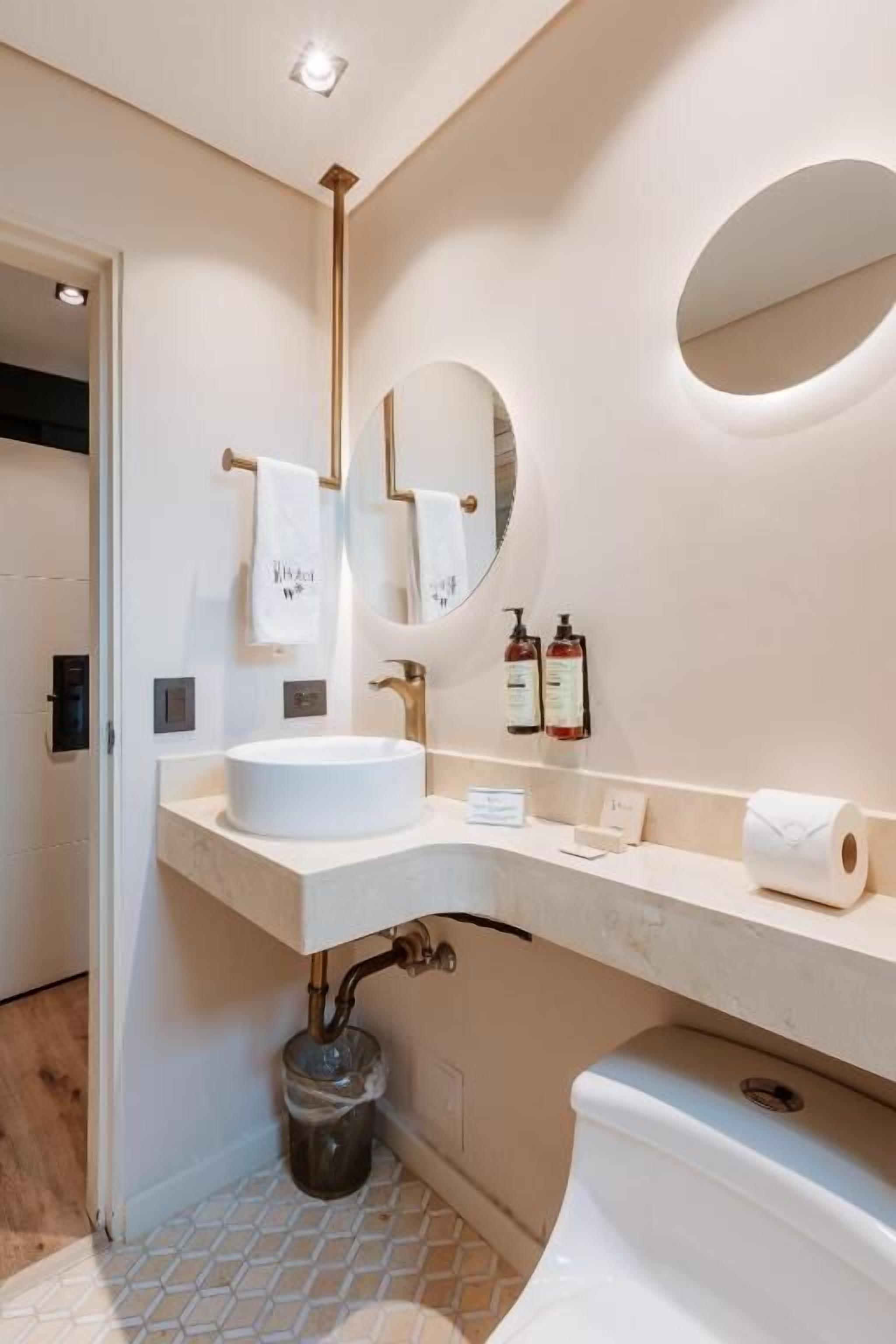 A bathroom with a white sink, mirror, and toilet. There is a towel rack with two towels, a trash bin, and a toilet paper roll.