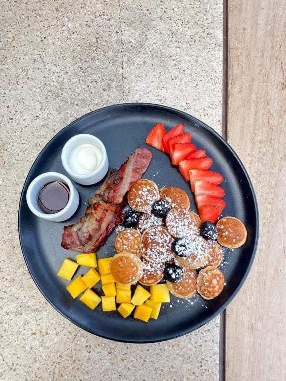 A breakfast plate with pancakes, bacon, strawberries, blueberries, and a side of fruit.