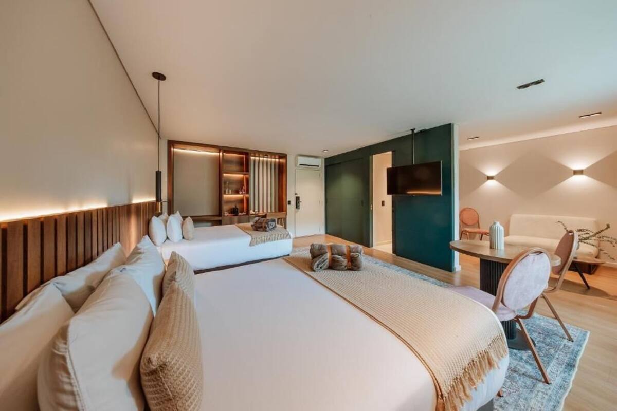 A modern hotel room with two beds, a TV, a dining area, and a large mirror.
