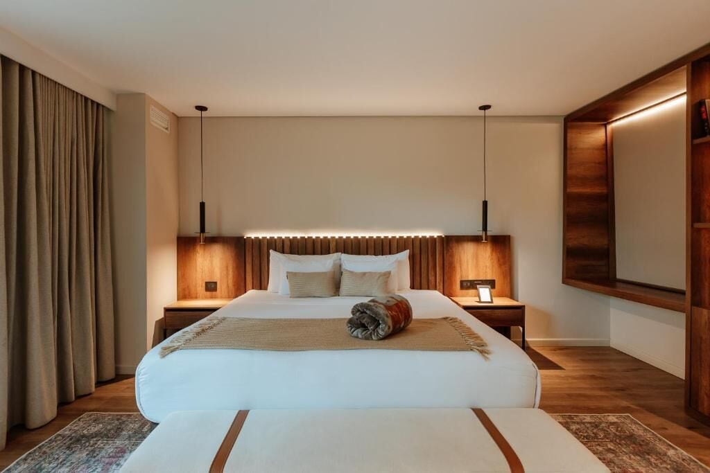A modern hotel room with a large bed, wooden headboard, bedside tables, and a flat-screen TV mounted on the wall.