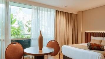 A hotel room with a bed, a round table with chairs, a vase, and a view of greenery outside.