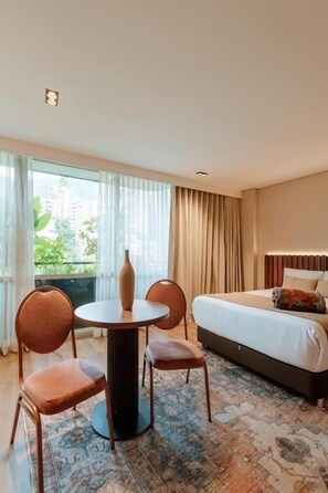 A hotel room with a bed, a round table with chairs, a vase, and a view of greenery outside.