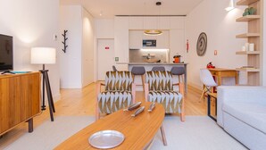 Een moderne woonkamer met een houten eettafel, twee gestreepte fauteuils, een bank, een tv op een houten kast, en een keuken op de achtergrond.
