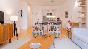 A modern living room with a wooden dining table, two patterned armchairs, a sofa, a TV on a wooden cabinet, and a kitchen in the background.