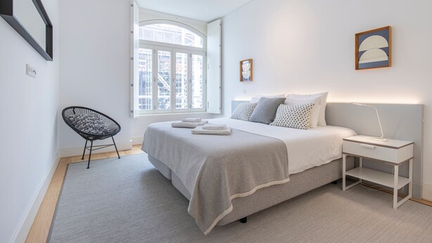 Een moderne slaapkamer met een groot bed, een nachtkastje met een lamp, een stoel en een raam met uitzicht op gebouwen.