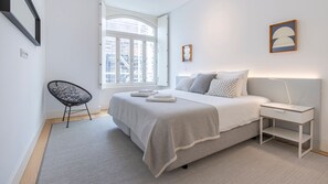 A modern bedroom with a large bed, a bedside table with a lamp, a chair, and a window with a view of buildings. - Spacious 1BDR in Santos by LovelyStay (Lisboa)