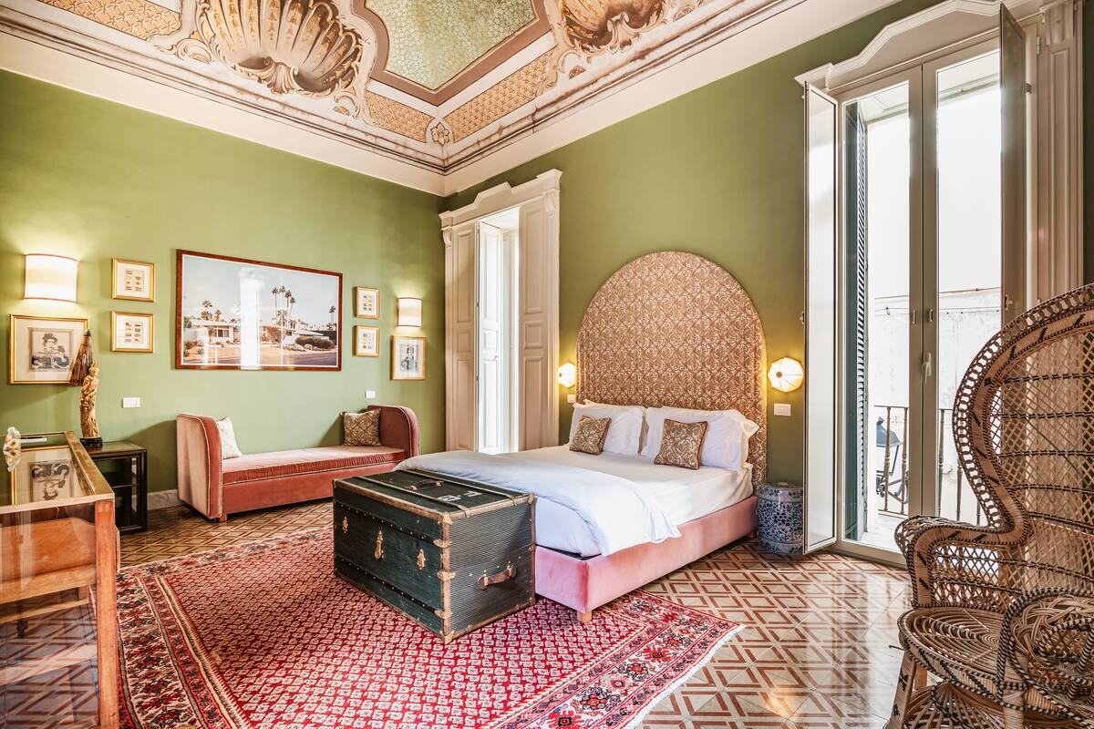 A bedroom with a large bed, a vintage trunk, a sofa, and a wicker chair.