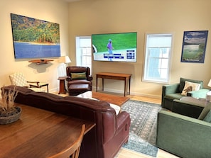 A living room with a wooden dining table, leather sofa, green armchair, and a flat-screen TV displaying a golf game.