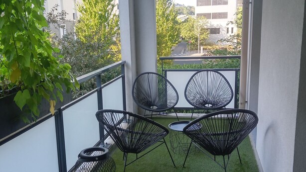 Ein Balkon mit einem Holzfass, zwei schwarzen Stuhlen aus Draht und einem kleinen Tisch.