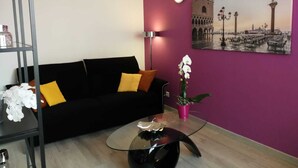 Ein Wohnzimmer mit einer schwarzen Couch, einem Glastisch, einem rosafarbenen Topfpflanzen und einer Wandlampe.