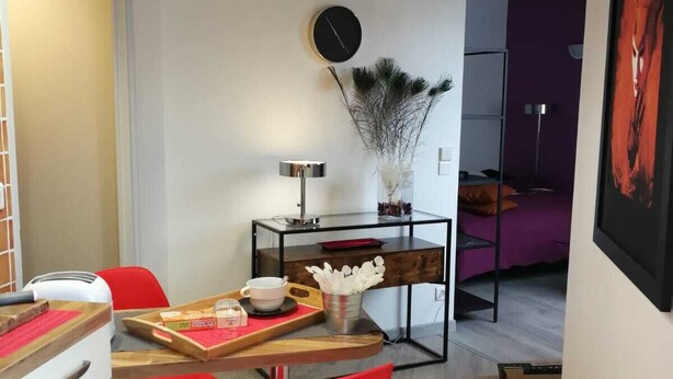 Ein kompakter Wohnbereich mit Essbereich, ausgestattet mit einem Holztisch, roten Stühlen und einer weißen Kommode. Auf einem Beistelltisch stehen eine moderne Lampe und eine dekorative Vase. Die Wand hat einen lila Farbakzent, und ein gerahmtes Kunstwerk ist ebenfalls zu sehen.