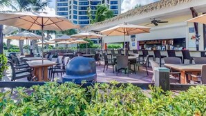 Un espace extérieur aménagé pour les repas, avec des parasols, des tables et des chaises, donnant sur une piscine et un bâtiment dont le toit est recouvert de chaume.