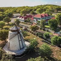 A windmill surrounded by a landscaped garden, a large house with a red roof, and a road in the background.