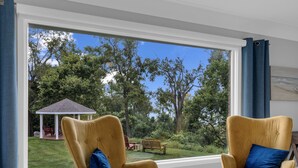 A room with two mustard-colored armchairs with blue cushions, a small white table, and a large window offering a view of a garden with a gazebo. - Coastal Suite #1 - Seacliff Beach Suites (Leamington)