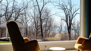 A room with a large window overlooking a forested area. There are two chairs and a small table in the room. - Coastal Suite #1 - Seacliff Beach Suites (Leamington)