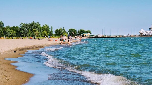 A beach with people walking along the shore, a clear sky, and a large building in the background. - Coastal Suite #1 - Seacliff Beach Suites (Leamington)