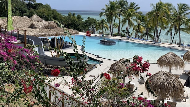 Zona de piscina en un resort con vista al océano, rodeada de palmeras y sombrillas de paja.