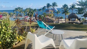 Terraza de madera con sillas de plástico blancas y una pequeña mesa, con vistas a una zona de piscina con palmeras y una estructura de techo de paja.