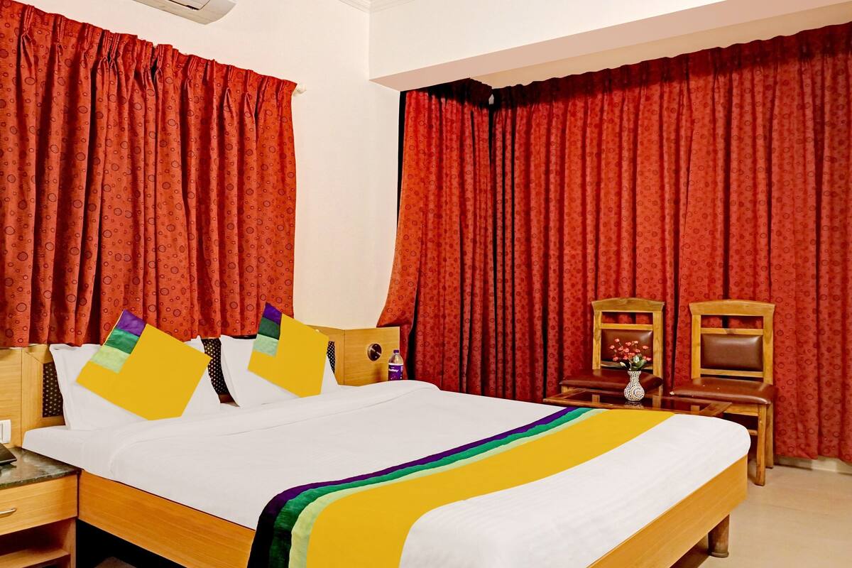 A hotel room with a bed, a wooden nightstand, a small table with a vase, and red curtains.