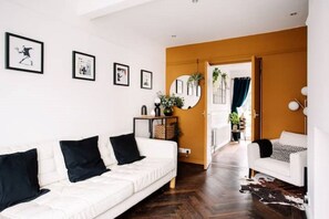 Ein modernes Wohnzimmer mit einem weißen Sofa, schwarzen Kissen, einem Couchtisch und einem Kuhfellteppich. An den Wänden hängen gerahmte Bilder und ein Durchgang zu einem weiteren Raum.