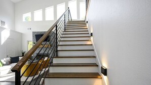 A modern staircase with wooden steps and a metal railing, leading to a living area with a fireplace. - Champagne Pines Escape | Hot Tub + EV | Sleeps 10 (Broken Bow)
