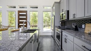A modern kitchen with white cabinets, granite countertops, a built-in microwave, and a large window overlooking a deck. - Champagne Pines Escape | Hot Tub + EV | Sleeps 10 (Broken Bow)