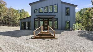 A modern two-story building with a central entrance, a gravel driveway, and a wooden staircase leading to the entrance. - Champagne Pines Escape | Hot Tub + EV | Sleeps 10 (Broken Bow)