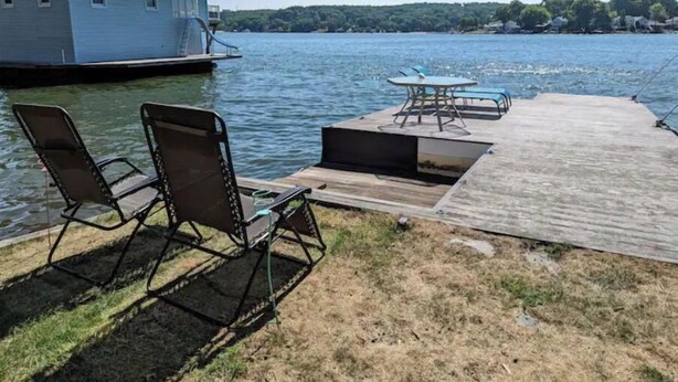Une jetée avec des chaises et une table au bord d’un lac, un petit bâtiment sur l’eau et, au loin, une colline boisée.