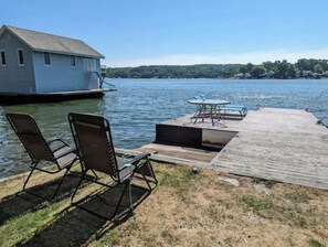 A dock with chairs and a table by a lake, a small building on the water, and a forested hill in the background. - Luxury 2BR Lakefront w/ Dock & Kayaks (Hopatcong)