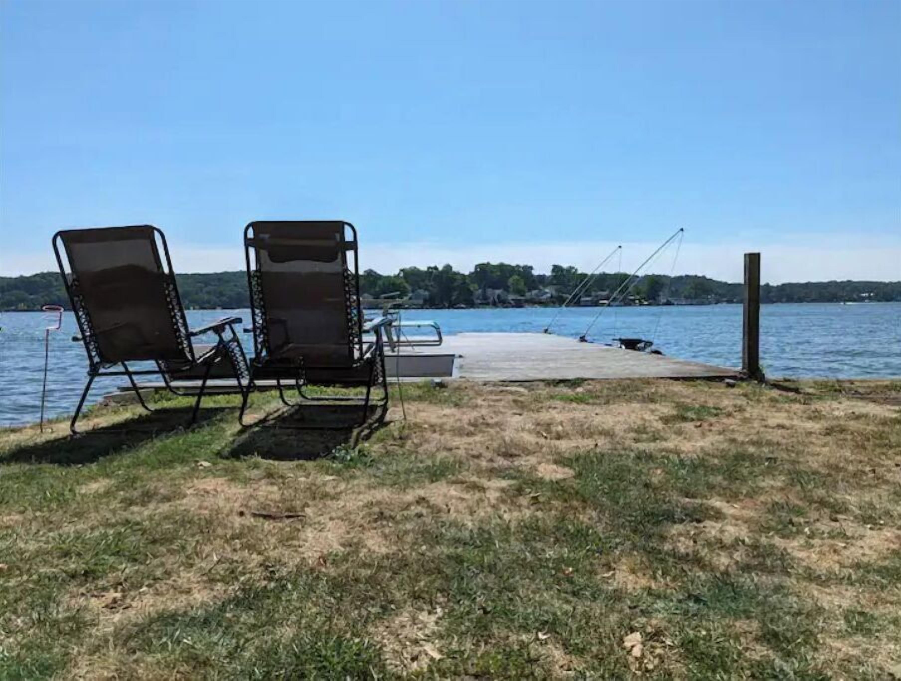 Two lawn chairs facing a dock by a lake with boats and fishing rods.