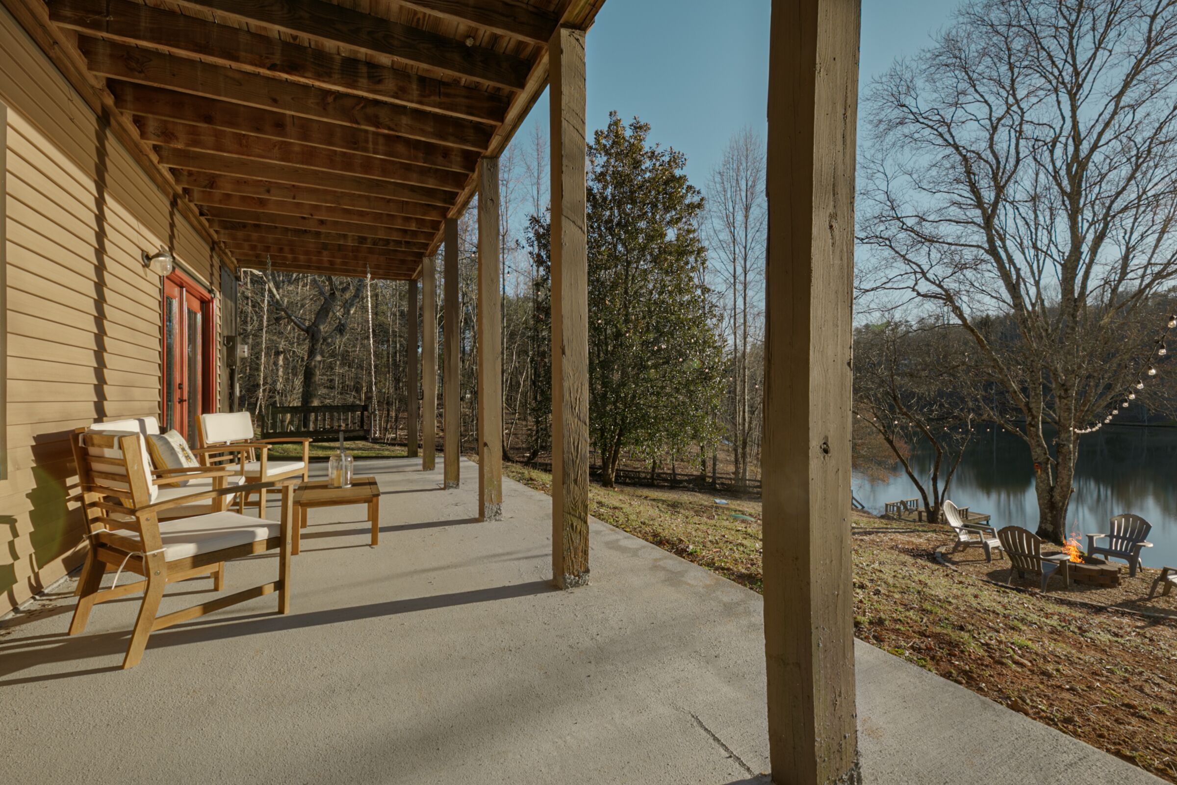 A covered outdoor area with wooden furniture, a concrete floor, and a view of a lake with trees and chairs.