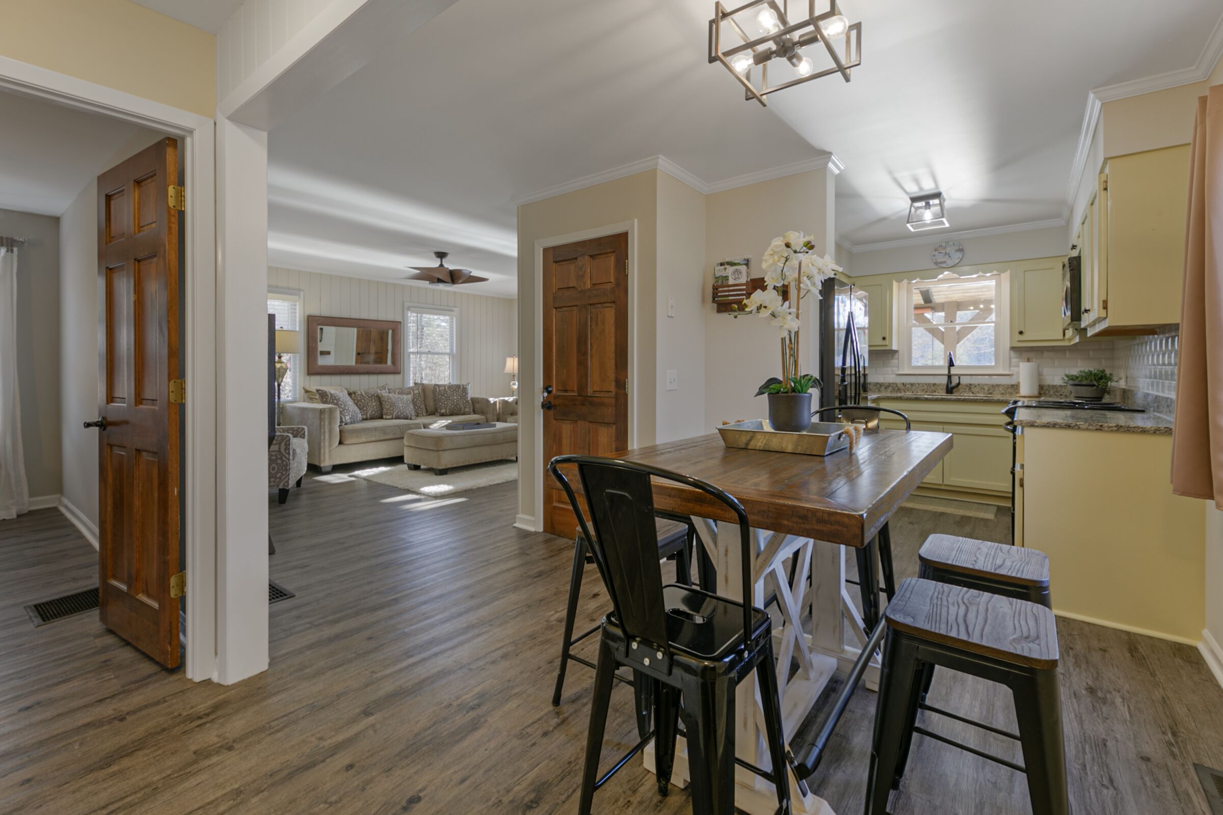 A spacious living room with a wooden dining table, black chairs, and a sofa. There is a kitchen area with a window and a refrigerator.