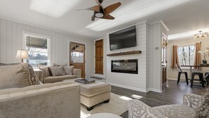 A spacious living room with a large TV mounted on the wall, a ceiling fan, and a fireplace. - Wine Country Lake Escape-Fishing and Hot tub (Cleveland)