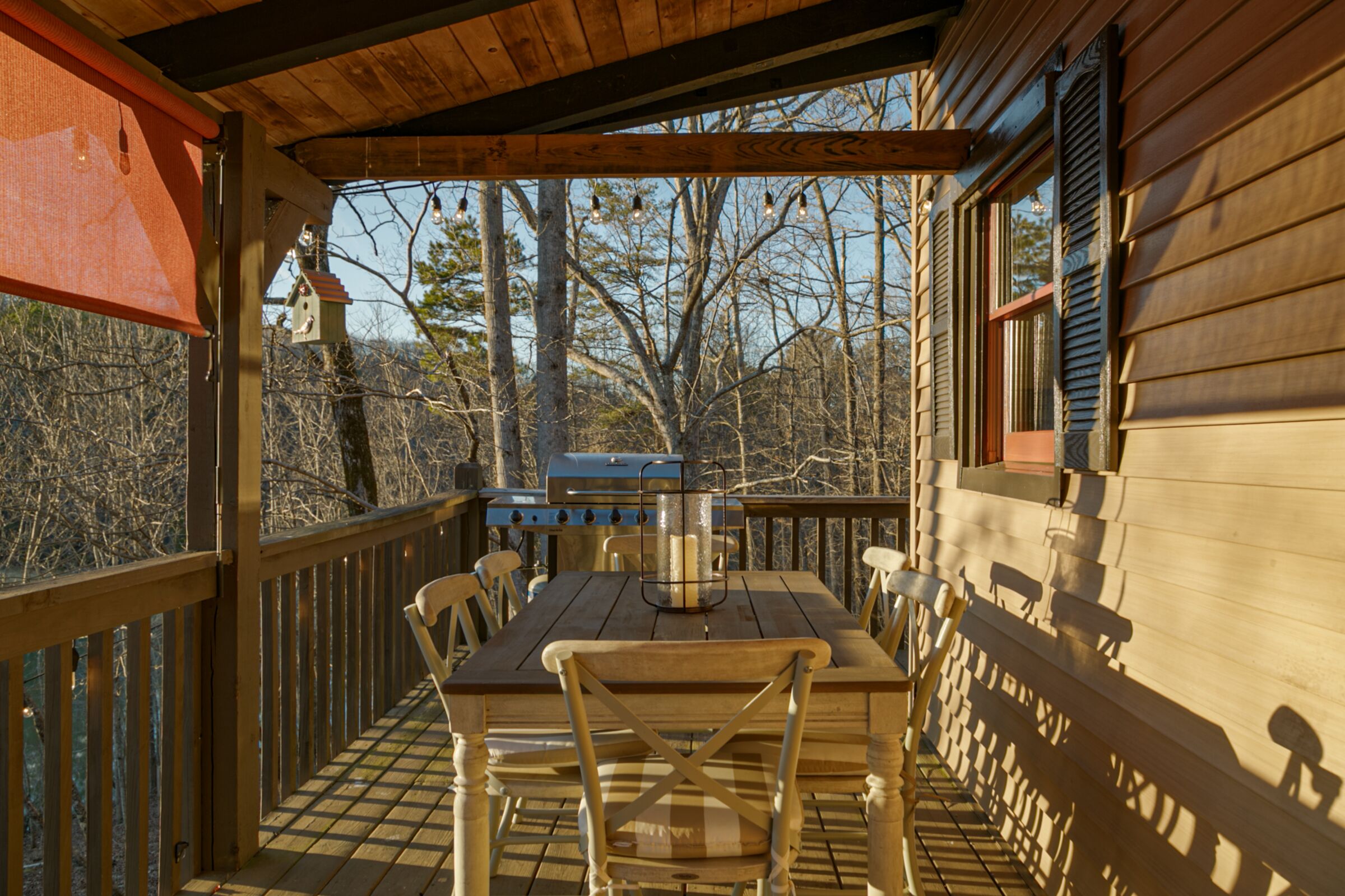An outdoor wooden deck with a table and chairs, a grill, and a view of trees and a birdhouse in the background.