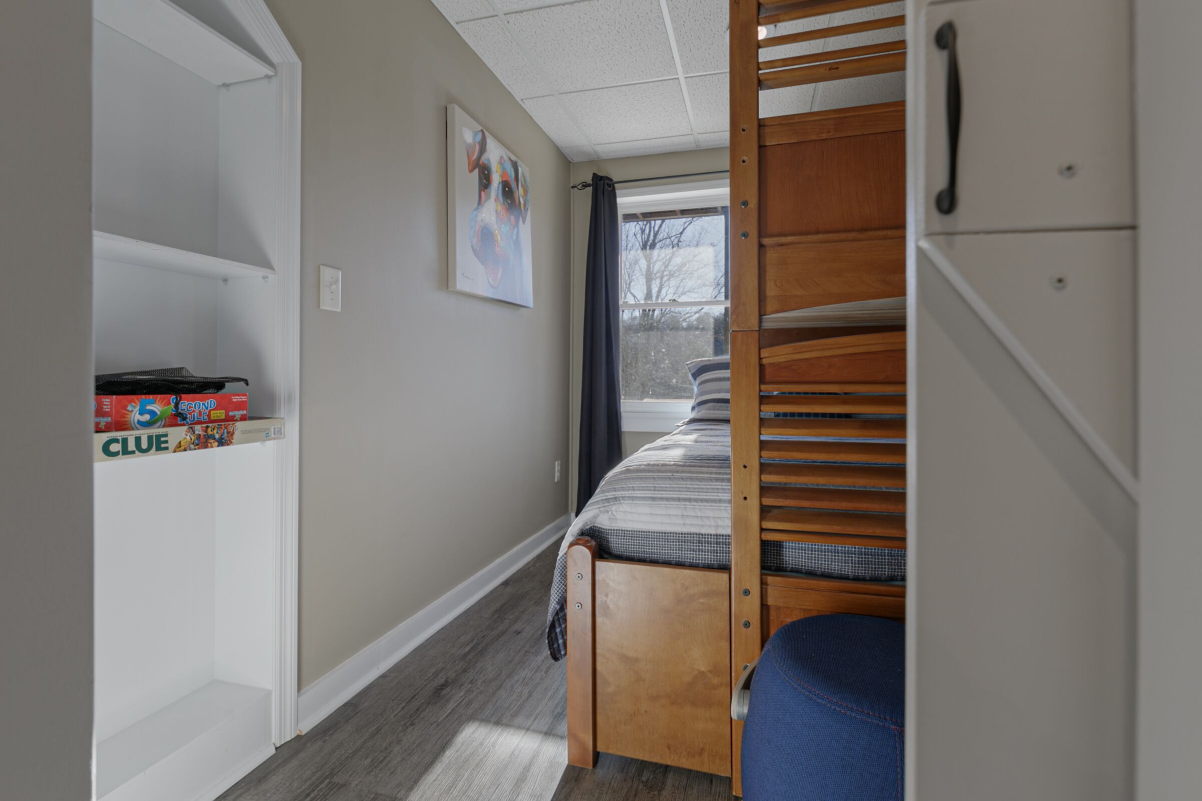 A dormitory room with a bunk bed, a shelf with storage boxes, a window with a view, and a door leading to another room.