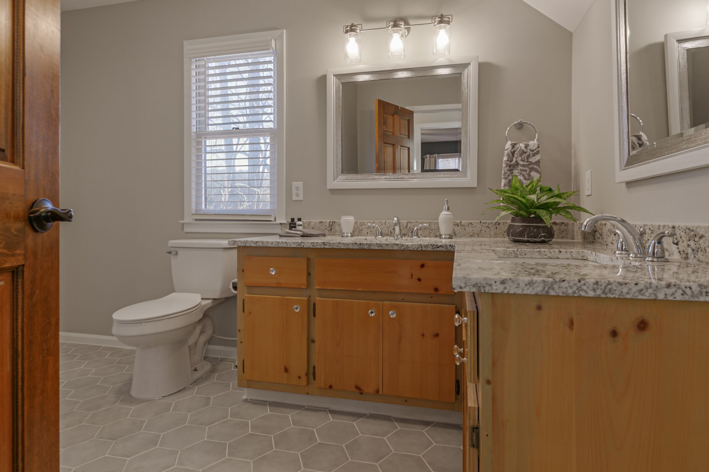 A bathroom with a toilet, wooden cabinets, a granite countertop, a sink, a mirror, and a window with blinds.