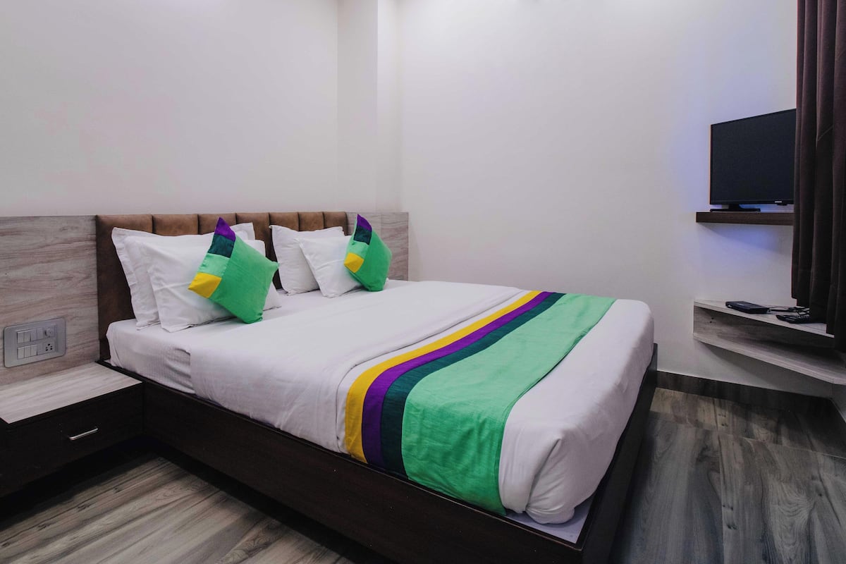 A neatly made bed with a white and multicolored striped blanket, a wooden headboard, and a television on a shelf to the right.